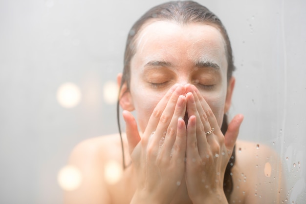 Close-up portrait of a woman with soap on her wet face standing behind the glass in the shower. Image with soft focus