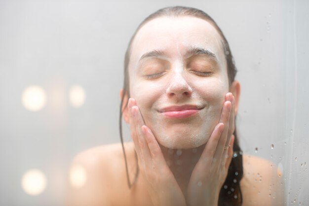 Close-up portrait of a woman with soap on her wet face standing behind the glass in the shower. Image with soft focus