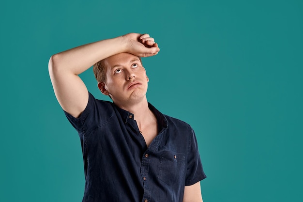 Close-up portrait of a young athletic ginger peson in a stylish navy t-shirt looking tired while posing on blue studio background. Human facial expressions. Sincere emotions concept. Copy space.