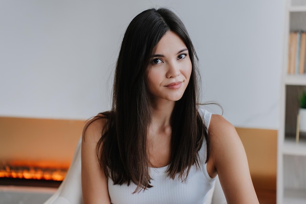 Photo close up portrait of young attractive asian woman with loose long hair looking at camera sits in chair over blurry fireplace at home pretty korean female in white tshirt beautiful models portrait