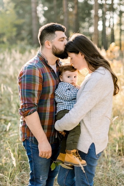 Close up portrait of young attractive family with little baby son, posing in beautiful autumn pine forest at sunny day