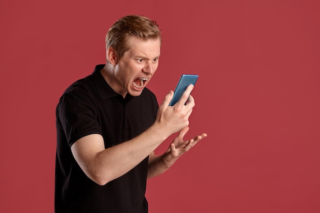 Close-up portrait of a young attractive ginger fellow in a stylish black t-shirt looking angry and shouting at his smartphone while posing on pink studio background. Human facial expressions. Sincere