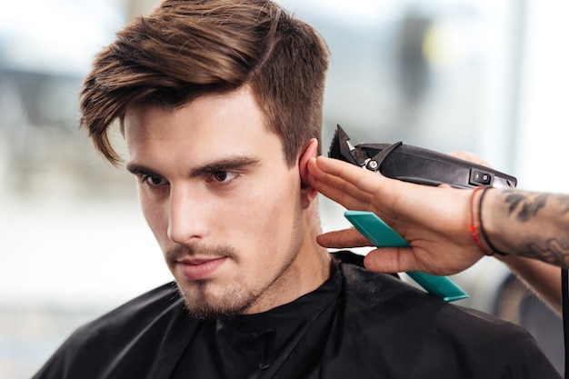 Close-up portrait of a young bearded man getting haircut by hairdresser while sitting in chair at barbershop