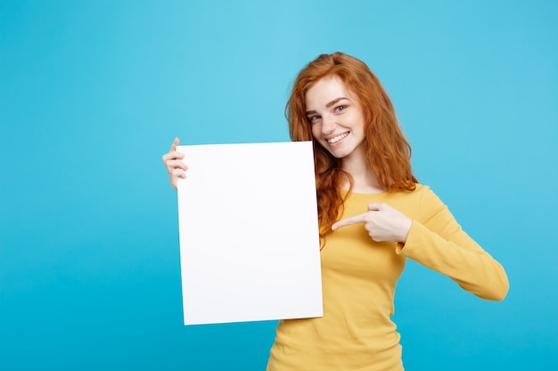 Close up Portrait young beautiful attractive ginger red hair girl smiling showing blank sign. Blue Pastel Background. Copy space.