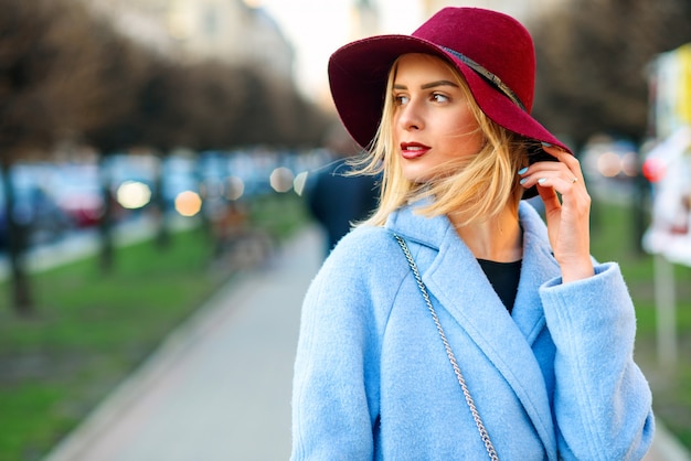 Close-up portrait of young beautiful girl in a blue coat and burgundy hat walking down the street on a sunny spring day