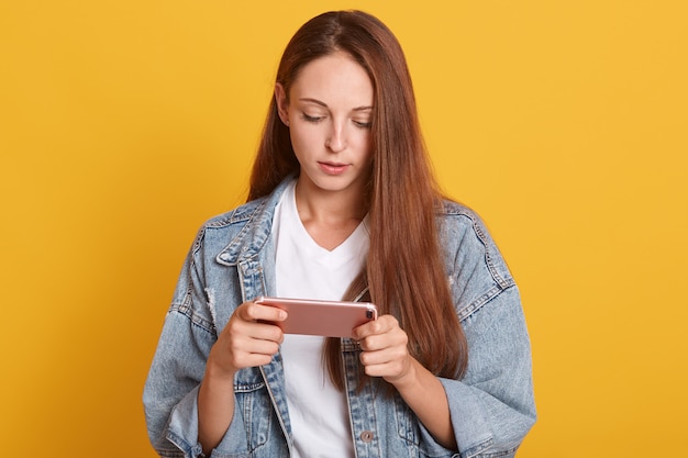 Close up portrait of young beautiful woman using smart phone, writing message to her friend, wearing denim jacket and white casual t-shirt, looking at device, looks concentrated.