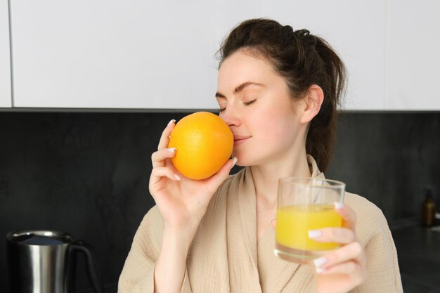 Close up portrait of young brunette smells fresh orange drinking juice from glass enjoying healthy