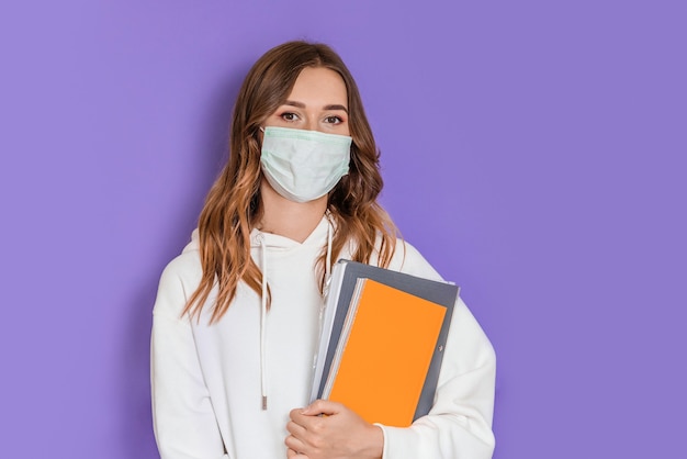 Close up portrait of young brunette student girl with surgical medical mask wearing a white hoodie standing and looking at camera isolated over lilac background