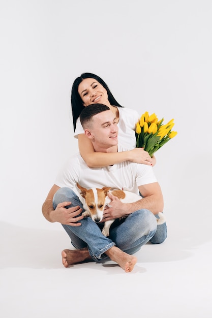 Close up portrait young couple sit and hugging, holding yellow flowers and dog in studio on white background. couple embracing with dreamy amorous expression. Lovely family. Celebrating woman's day.