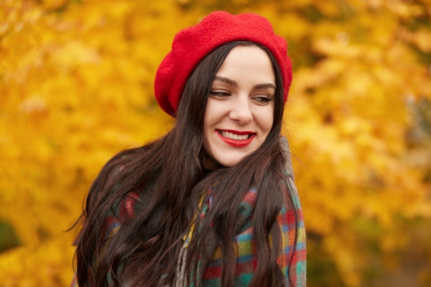 Close up portrait of young fashionable girl wears red beret, having dark long straight hair, resting in park in autumn stands around yellow trees