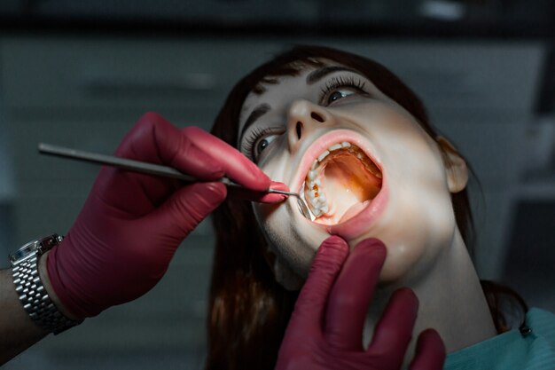 Close up portrait of young female patient with open mouth having professional dental treatment, and hands of doctor wearing red rubber gloves, holding dental mirror