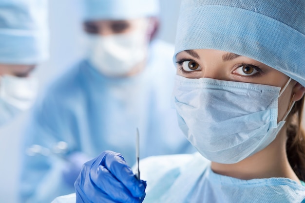 Close up portrait of young female surgeon doctor holding scalpel surrounded by her team