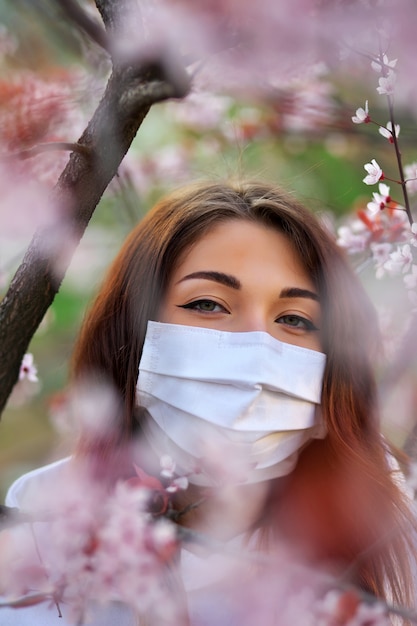 Foto chiuda sul ritratto della ragazza sotto un albero di albicocca sbocciante con una maschera dal coronavirus.