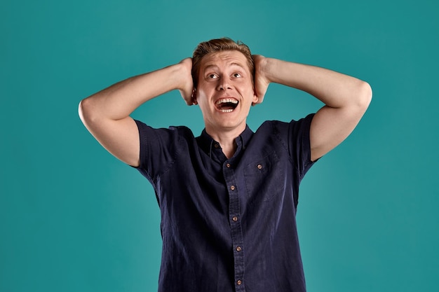 Close-up portrait of a young good-looking ginger guy in a stylish navy t-shirt looking shocked about something while posing on blue studio background. Human facial expressions. Sincere emotions concep