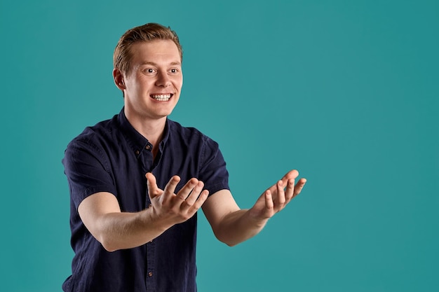 Close-up portrait of a young good-looking ginger male in a stylish navy t-shirt pulling hands to someone and smiling while posing on blue studio background. Human facial expressions. Sincere emotions