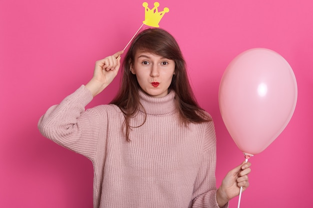 Close up portrait of young happy woman holding toy paper crown above her head and pink balloon.