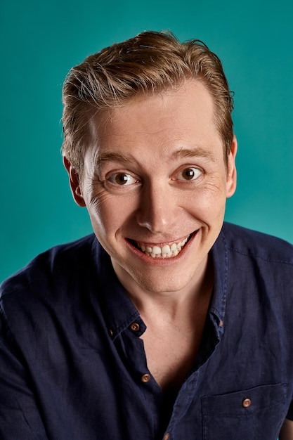 Close-up portrait of a young interesting ginger peson in a stylish navy t-shirt looking at the camera and laughing while posing on blue studio background. Human facial expressions. Sincere emotions co