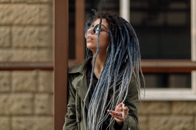 Photo close up portrait of a young pretty smiling woman with dreadlocks, cheerfully looking at the camera walking in city. girl smokes a cigarette