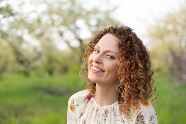 Close up portrait of young smiling attractive woman with curly hair in green flowering spring park. Pure emotions.