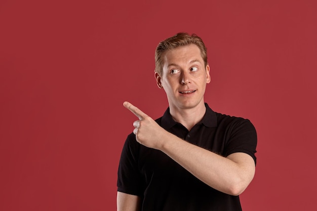 Close-up portrait of a young stately ginger fellow in a stylish black t-shirt pointing at something while posing on pink studio background. Human facial expressions. Sincere emotions concept. Copy spa