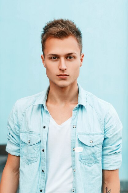 Close up portrait of a young stylish guy in a denim shirt near a blue wall