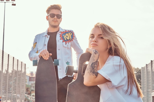 Close-up portrait of young trendy dressed hipster couple with skateboards posing at city sports complex on sunny day, with warm toned.