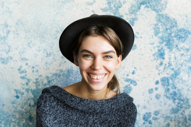 Close up portrait of a young woman dressed in sweater and hat over blue wall, laughing