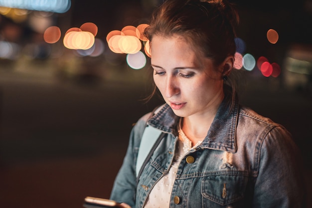 Ritratto del primo piano di una giovane donna che esamina il suo smartphone la notte