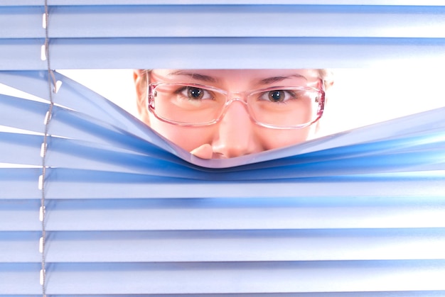 Close-up portrait of young woman looking through window blinds