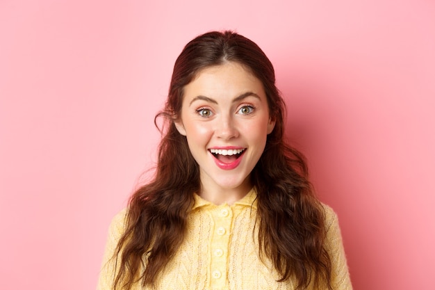 Close up portrait of young woman open mouth and gasping amazed, hear surprise news, looking impressed and happy, standing against pink wall.