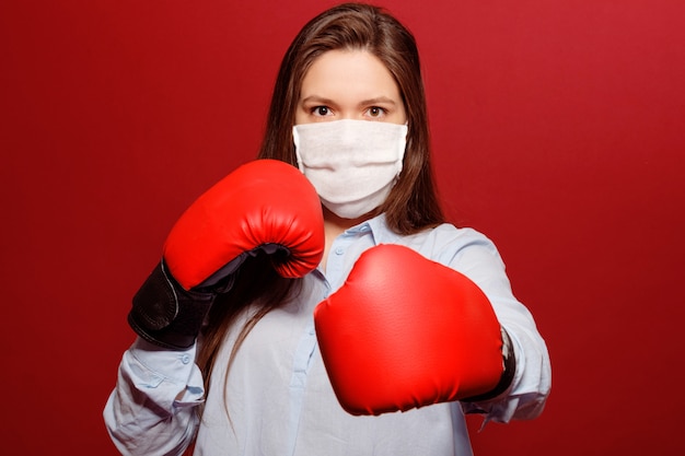 Close-up portrait of young woman in red boxing gloves on red background in protective medical mask, coronavirus pandemic, fight with virus