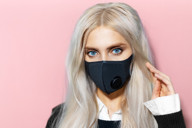 Close-up portrait of young woman wearing respiratory mask against coronavirus on background of pastel pink color.