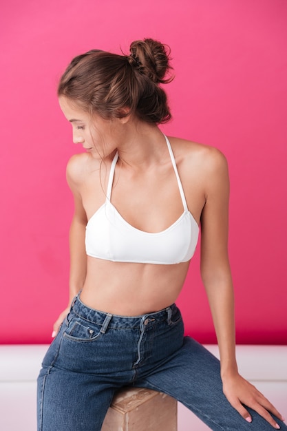 Close up portrait of a young woman in white top and jeans looking away isolated on the pink wall