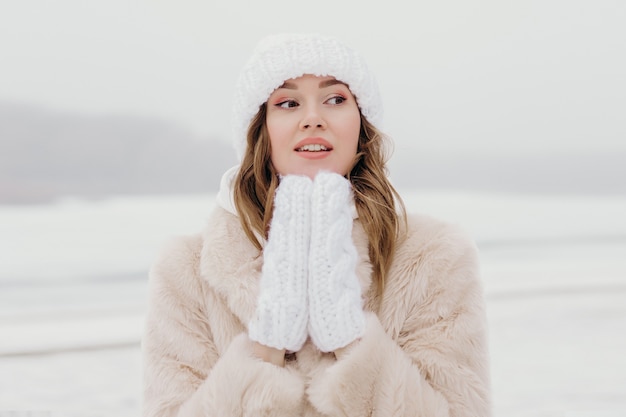 Close up portrait of a young woman in a winter hat and white gloves blowing snow and warming her hand.