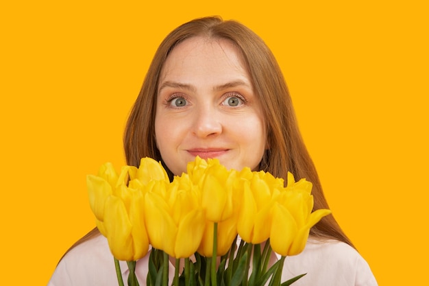 Close up portrait of young woman with gray eyes with bouquet of yellow tulips flowers isolated on yellow background