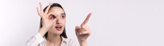 Close-up portret van aantrekkelijke eigenzinnige jonge vrouw die een verrekijker maakt met handen die ok gebaar op witte studio-achtergrond tonen