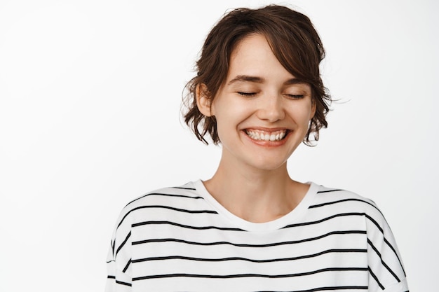 Close-up portret van aantrekkelijke gelukkige vrouw, glimlachend met gesloten ogen, romantisch natuurlijk meisje met gloeiende schone gezichtshuid, staande tegen een witte achtergrond