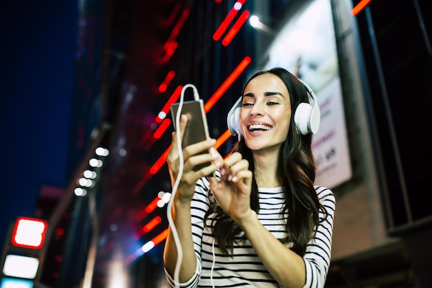 Close-up portret van aantrekkelijke jonge gelukkige vrouw in koptelefoon met slimme telefoon in handen buitenshuis