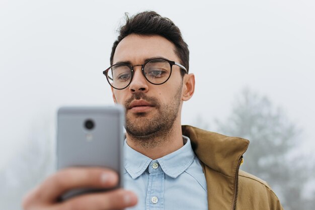 Close-up portret van blanke man dragen bril en jas wandelen buiten in mistig weer messaging via sociale netwerken op slimme telefoon en met behulp van draadloze mensen lifestyle technologie concept