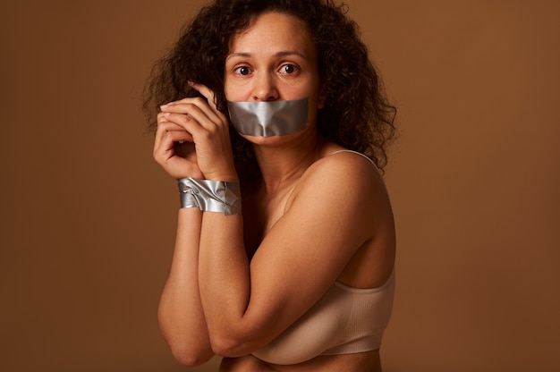 Close-up portret van een bange wanhopige vrouw met haar mond verzegeld en gebonden handen, kijkend naar de camera met hoop, smekend om hulp bij het beëindigen van geweld tegen vrouwen. Sociaal concept op donkere achtergrond