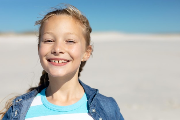 Close-up portret van een blank meisje op een zonnig strand, glimlachend naar de camera, met blauwe lucht en zand op de achtergrond