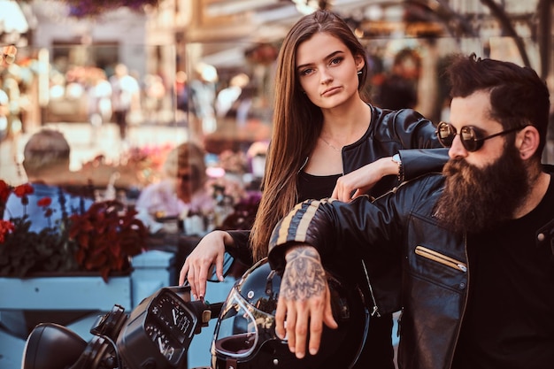 Close-up portret van een hipster paar - bebaarde brutale man in zonnebril gekleed in een zwart leren jas en zijn jonge sensuele meisje permanent in de buurt, poseren tegen terras van café.