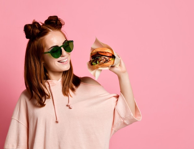 Close-up portret van een hongerige jonge vrouw die hamburger eet geïsoleerd over roze background