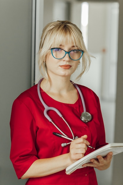 Close-up portret van een jonge blonde dokter in een rood uniform met een stethoscoop. Een dokter met een bril staat in het ziekenhuis, houdt een notitieboekje met een pen vast en kijkt naar de camera