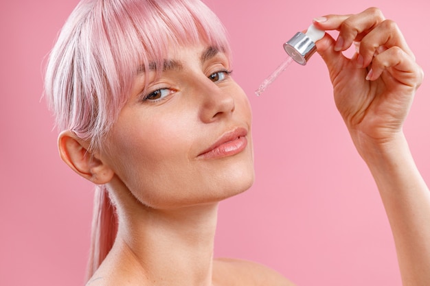 Close-up portret van een jonge vrouw met roze haar die serum of hyaluronzuur op haar gezichtshuid aanbrengt