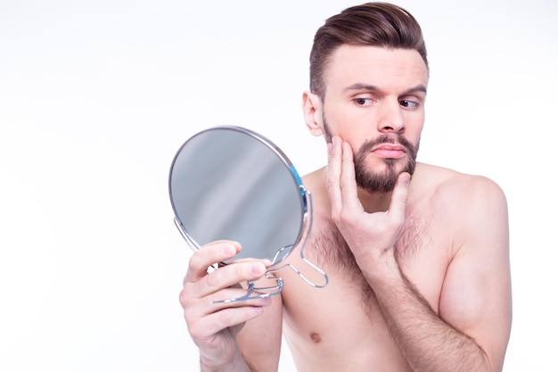 Close-up portret van een knappe bebaarde man met handdoek kijkend naar zijn spiegelbeeld in de spiegel in de badkamer geïsoleerd op een witte achtergrond