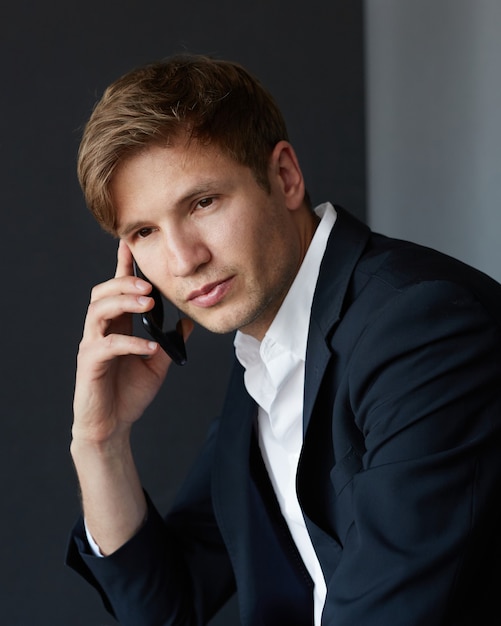 Close-up portret van een knappe jonge zakenman in een elegant pak die naar de camera zit te kijken, zijn telefoon bij zijn oor houdt, over een donkergrijze achtergrond.