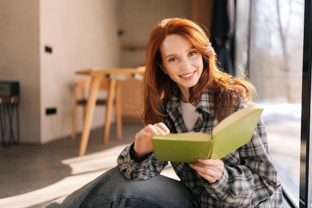 Close-up portret van een mooie vrouw met een leesboek die op een zonnige dag bij het raam zit en glimlacht