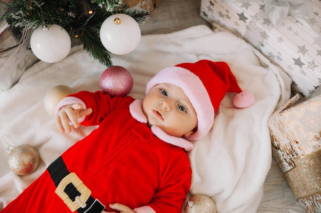 Close-up portret van een pasgeboren baby. Schattige blanke baby 5-6 maanden oud in kerstmankostuum liggend op een gezellige deken in de buurt van een versierde kerstboom. Vrolijk kerstfeest 2022