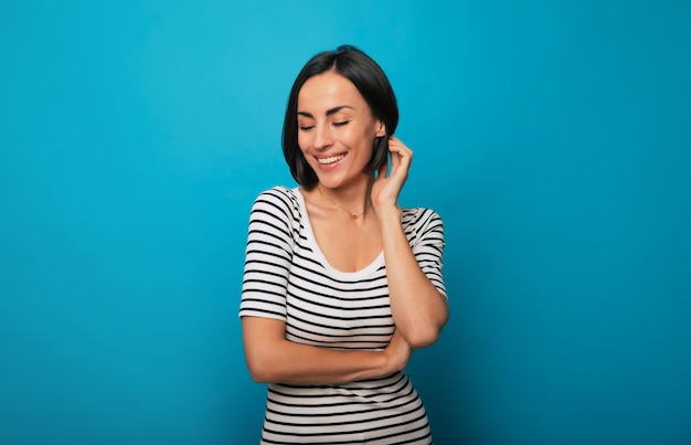 Close-up portret van een prachtige lachende jonge brunette vrouw terwijl ze poseren in een gestreepte t-shirt met gekruiste armen en geïsoleerd op blauwe achtergrond